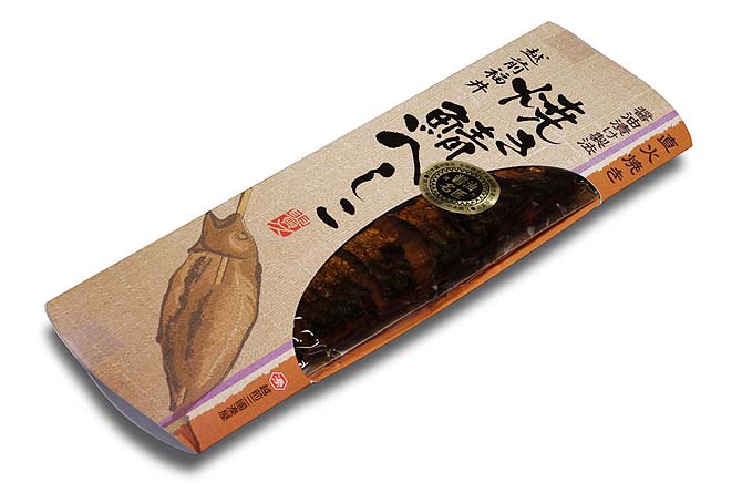 縦長筒状を押しつぶした構造。右側中央に中の商品が見える窓開き。[焼き鯖へしこ]の筆書き文字と一本串の焼き鯖イラスト。