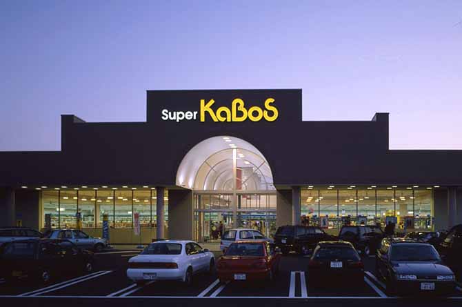 勝木書店SUPER KABOSの黄色と白のネオン文字が、建物が逆光で黒ずんだ壁面のシルエットとともに夕景の空に浮かんでいる。全景は駐車場の車が整然と並んで知る。